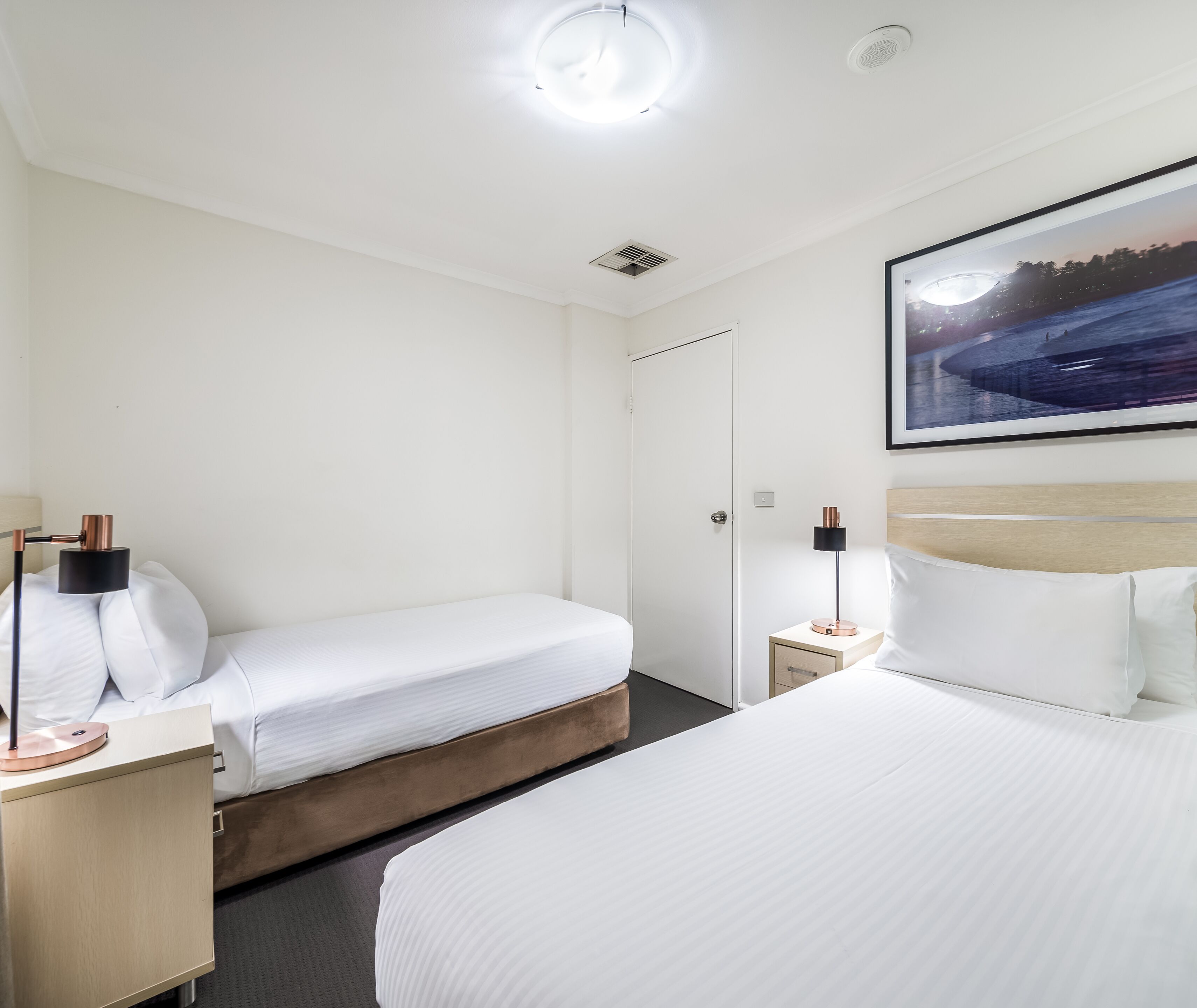 A hotel room with two beds, a nightstand with a lamp, a ceiling light, and a framed picture on the wall.