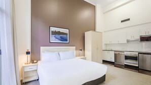A hotel room with a bed, a nightstand, a lamp, a picture on the wall, a kitchenette with a stove and microwave, and a cabinet. - Oaks Sydney Goldsbrough Suites (Pyrmont)
