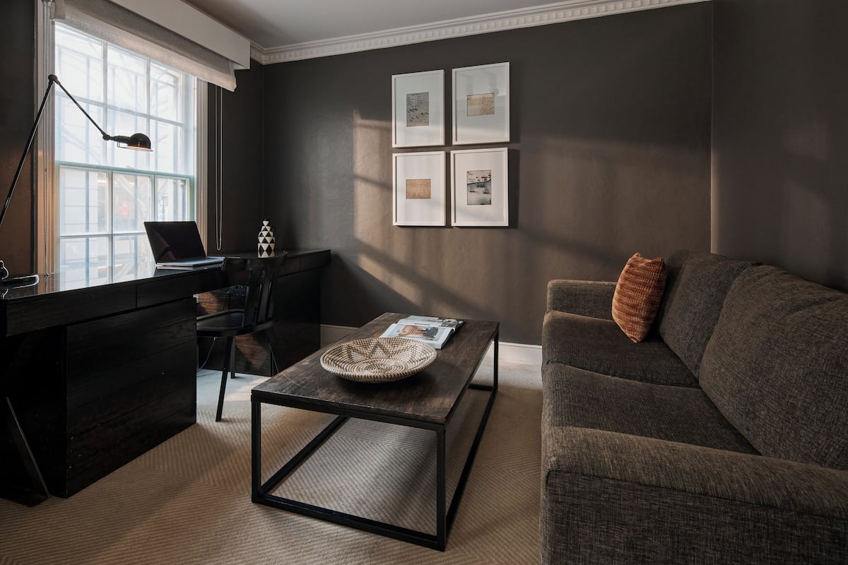A modern living room with a dark brown sofa, a coffee table, a desk with a laptop, and a large window.