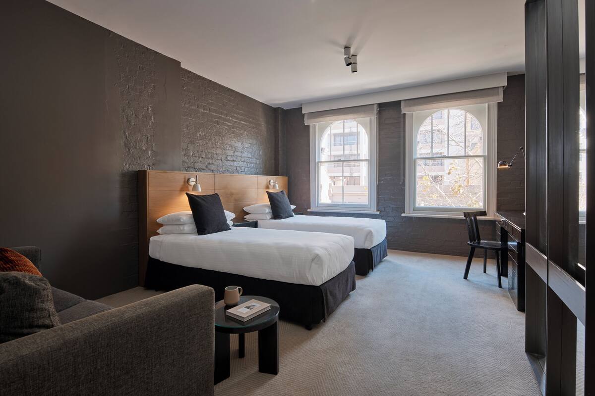 A modern hotel room with two beds, a sofa, a small table, and a large window providing a view of the city.