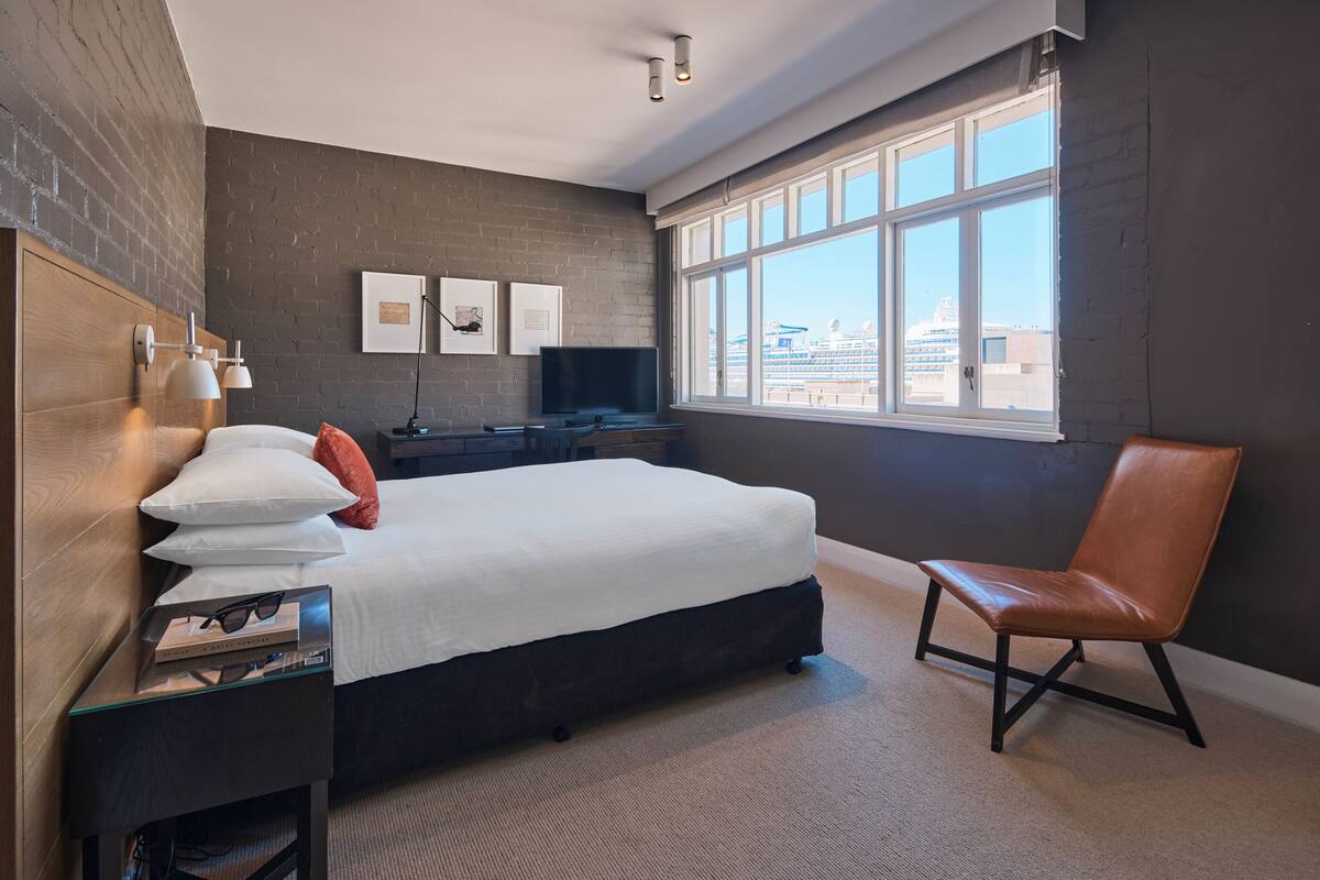 A modern hotel room with a large bed, a flat-screen TV, a window with a city view, and a leather chair.