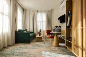 A hotel room with a green sofa, a small round table, a wooden wardrobe, and a TV mounted on the wall.