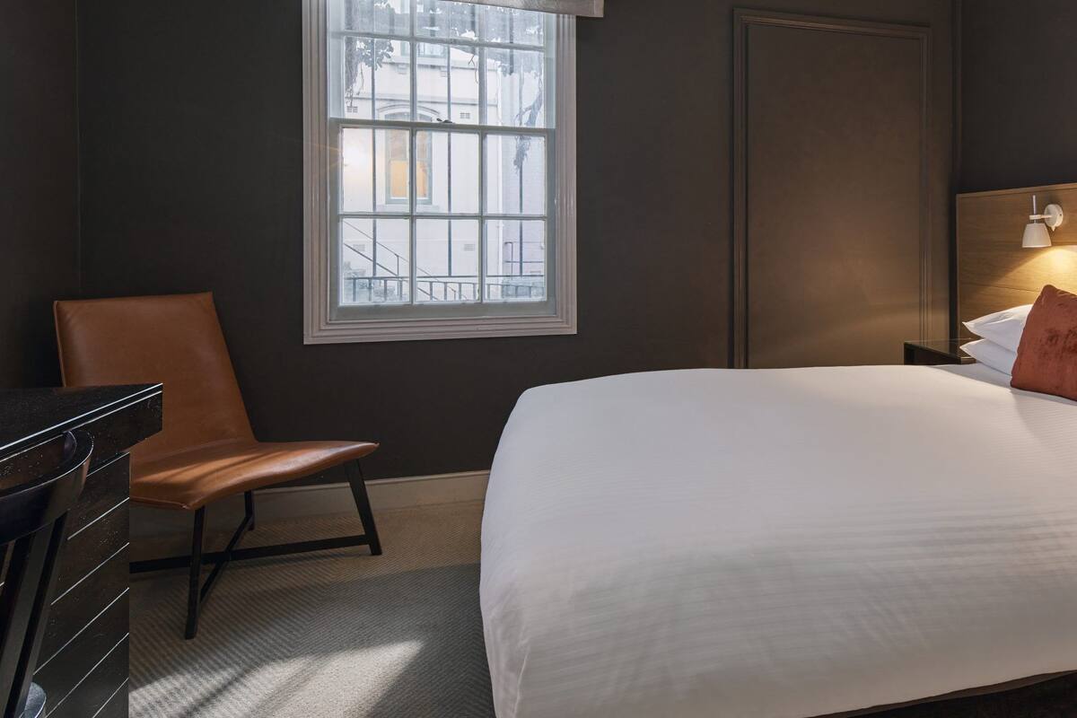 A hotel room with a large bed, a wooden headboard, a nightstand, a desk, a chair, and a window with a view of buildings.