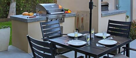 An outdoor kitchen with a grill, dining table, chairs, and a built-in oven.