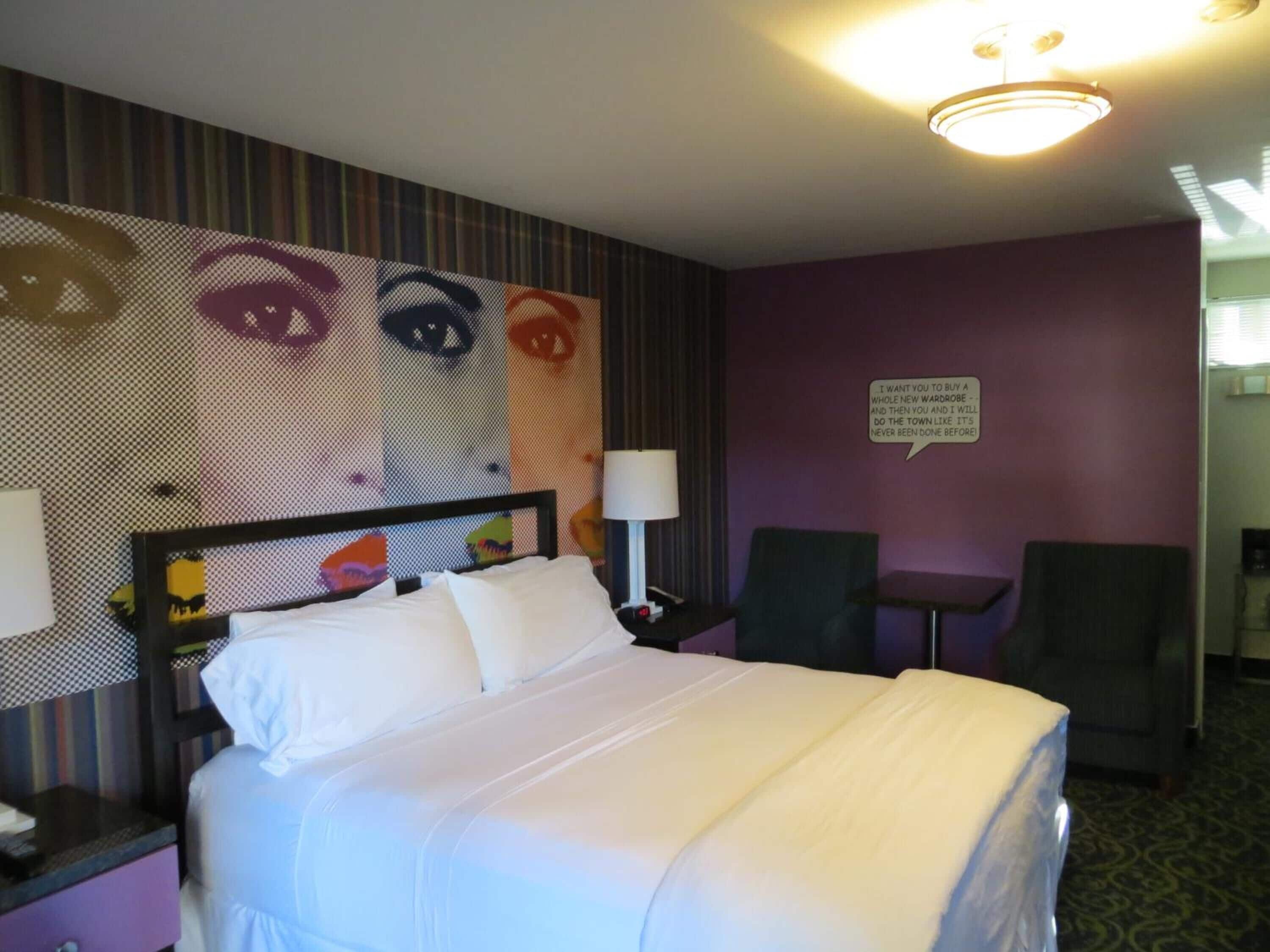 A hotel room with a bed, two chairs, a small table, and a wall with a graphic.