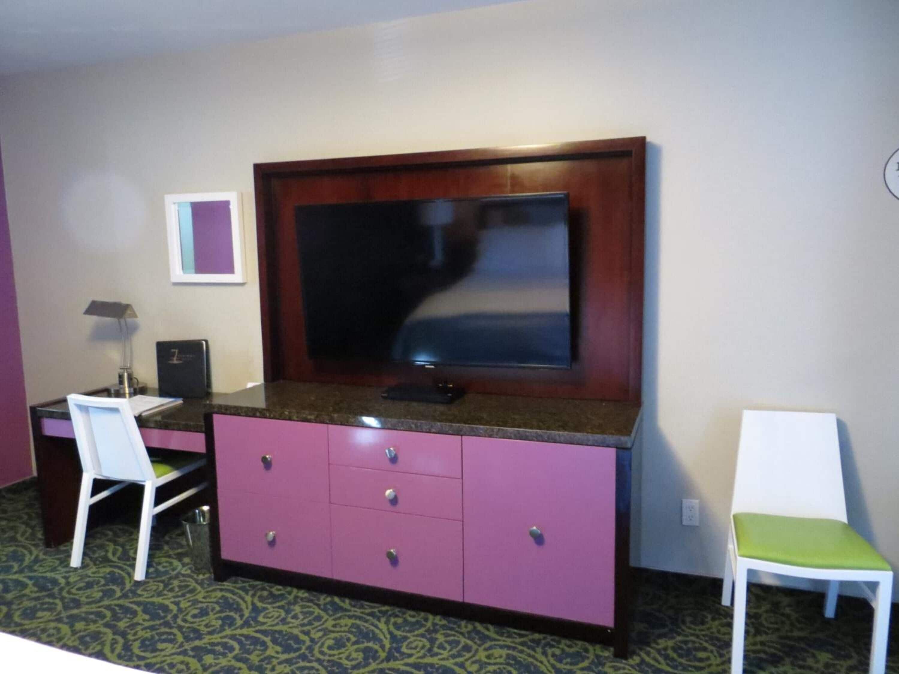 A hotel room with a large flat-screen TV mounted on a wooden cabinet, a desk with a chair, and a small table with a lamp.
