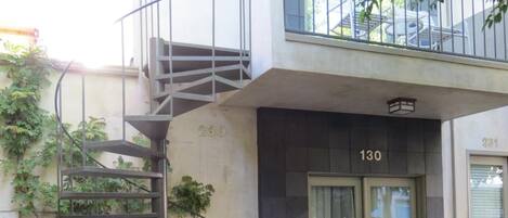 Un edificio moderno con una escalera que conduce a un balcón, una puerta con la etiqueta “130” y asientos al aire libre.