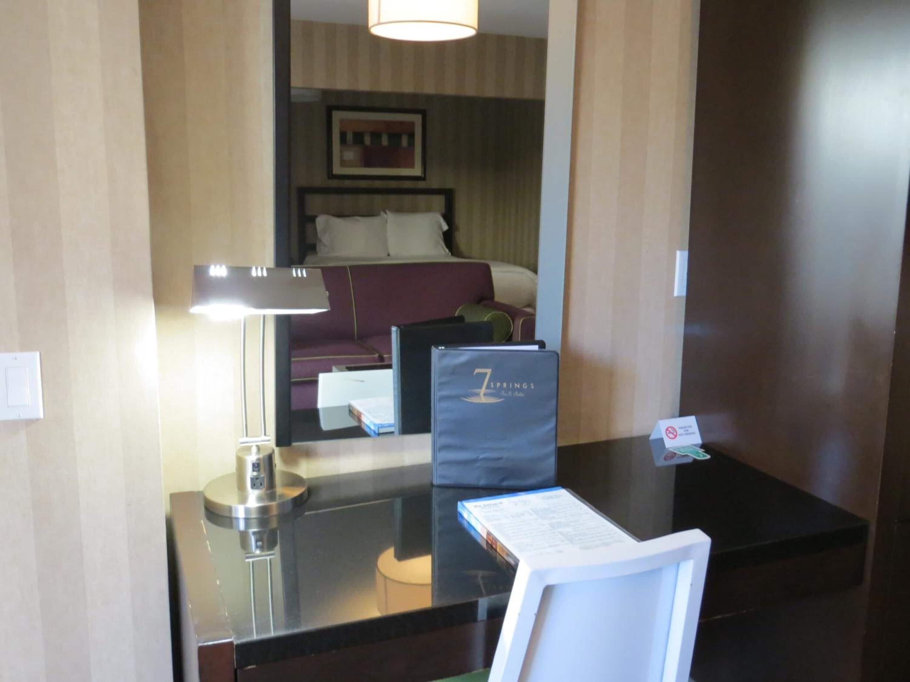 Balcão de recepção de hotel com uma cadeira de couro preto, uma luminária de mesa, uma pasta preta com a etiqueta '7 Springs' e uma moldura com uma fotografia na parede.