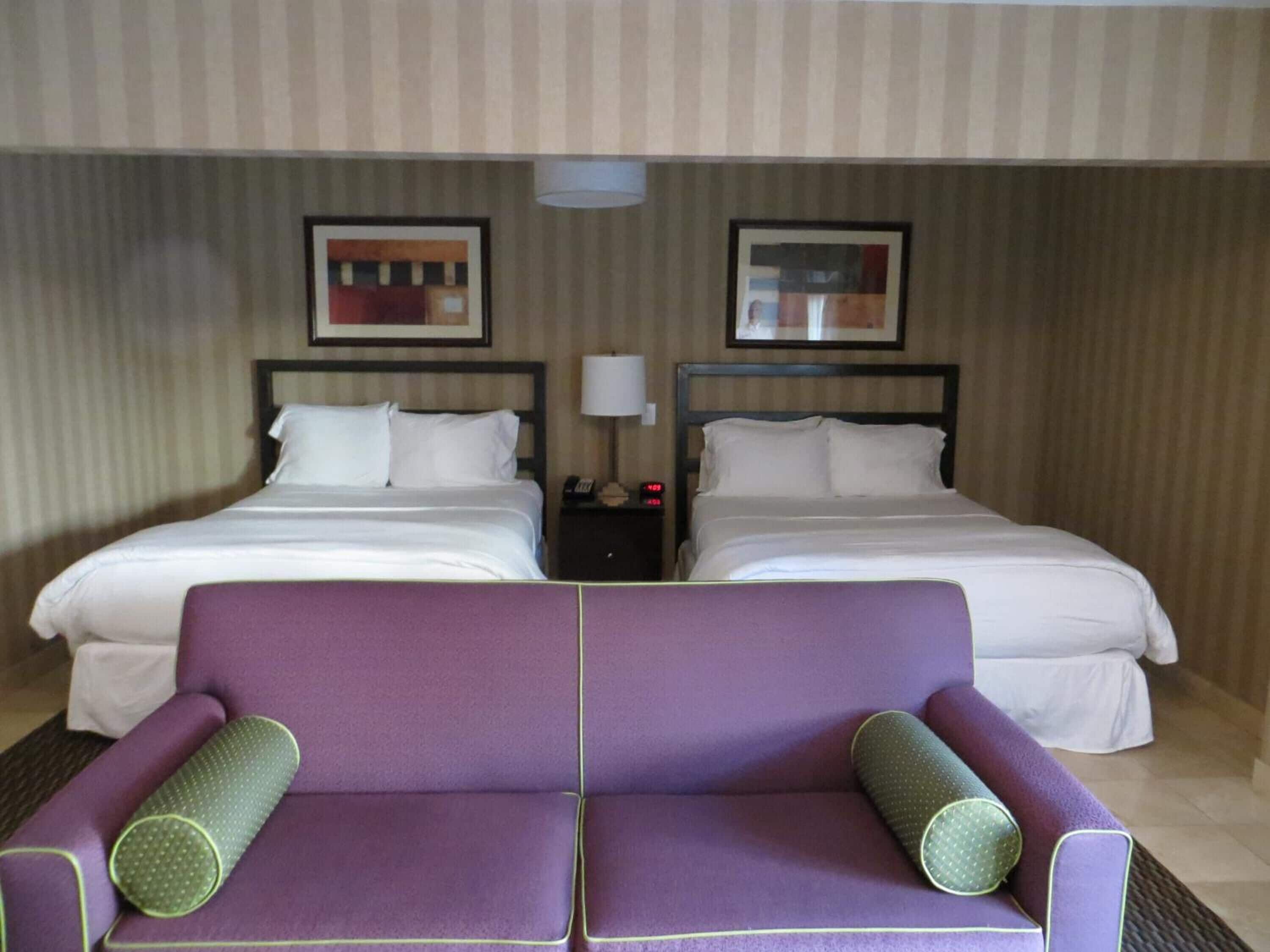 A hotel room with two beds, a nightstand with a lamp, and a purple sofa with two pillows.