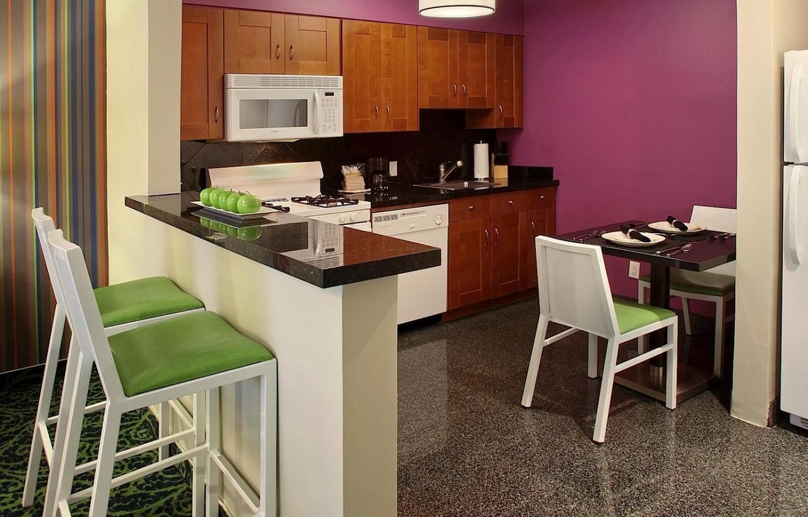A compact kitchen with a microwave, stove, and sink. There are two bar stools and a dining table with chairs.