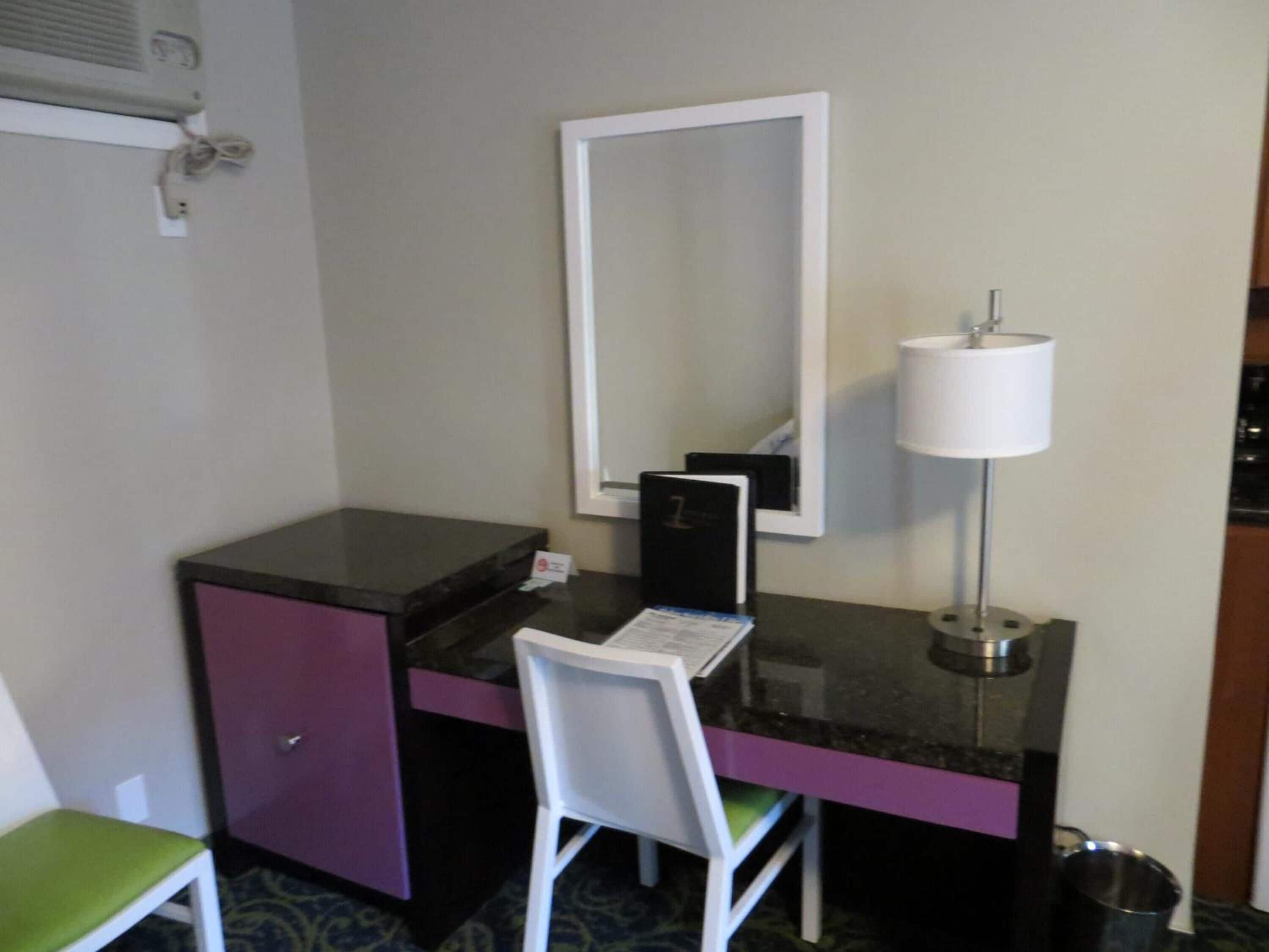 A small hotel room with a desk, chair, mirror, lamp, and a trash bin.