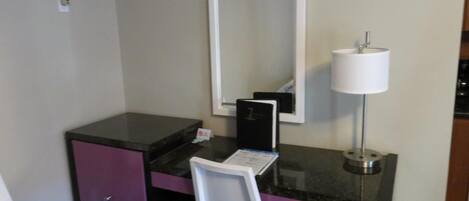 A small hotel room with a desk, chair, mirror, lamp, and a trash bin.
