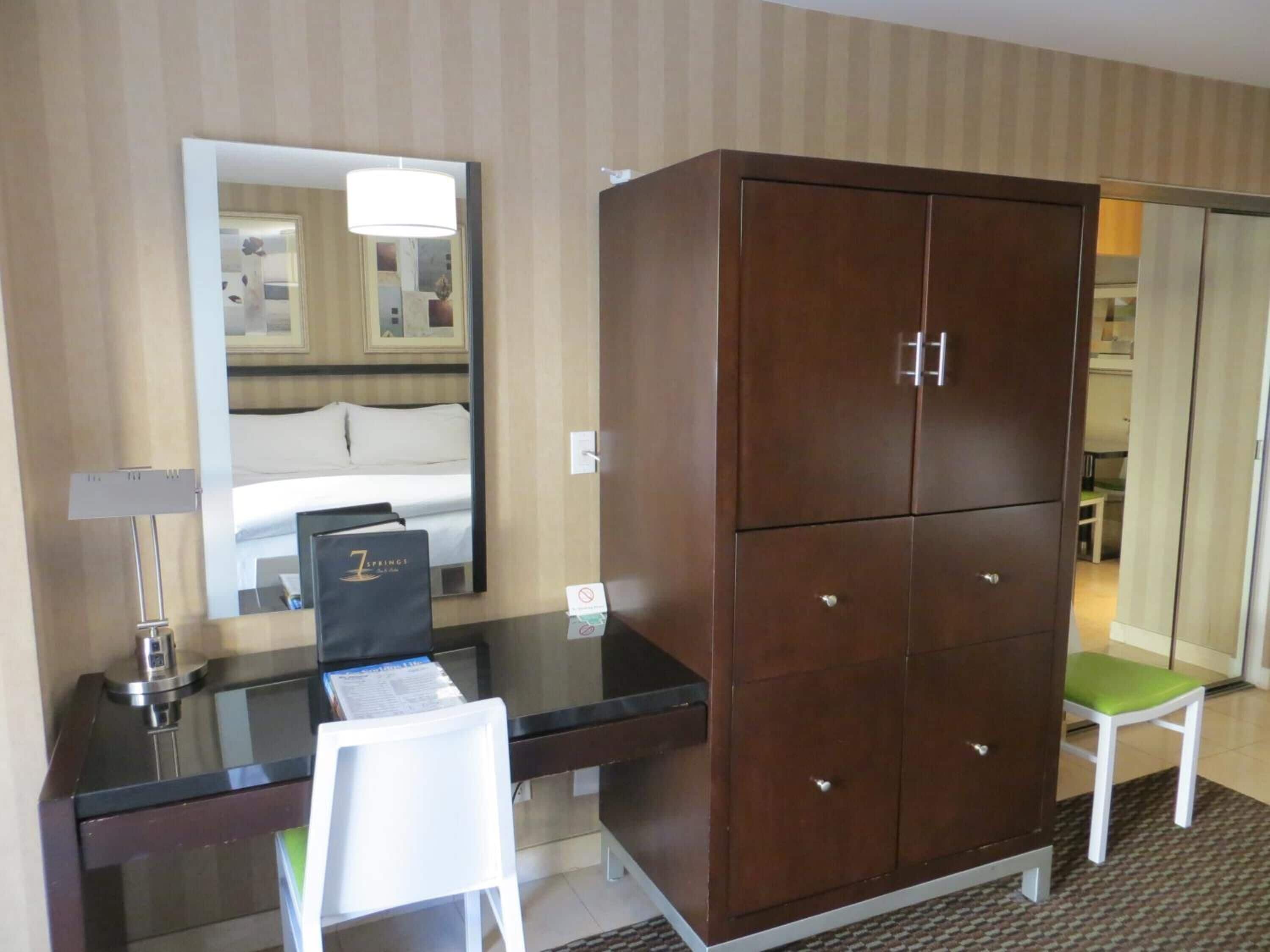 A hotel room with a large wooden wardrobe, a desk with a chair, a mirror, and a bed with white linens.