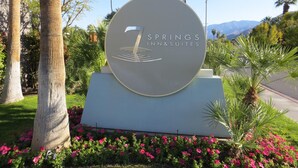 A sign for Springs Inn & Suites with a circular logo, surrounded by palm trees and flowering plants. - 7 Springs Inn & Suites (Palm Springs)