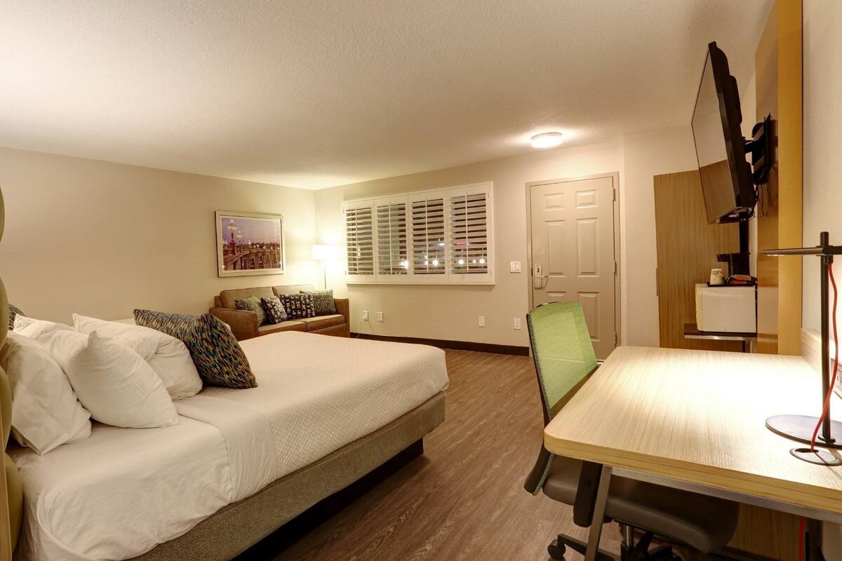 A hotel room with a bed, a desk, a chair, a lamp, a window with blinds, a door, and a wall-mounted TV.