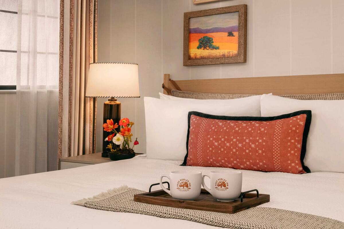 A neatly made bed with white linens, a wooden headboard, and decorative pillows. A tray with two white mugs and a cup is placed on the bed.
