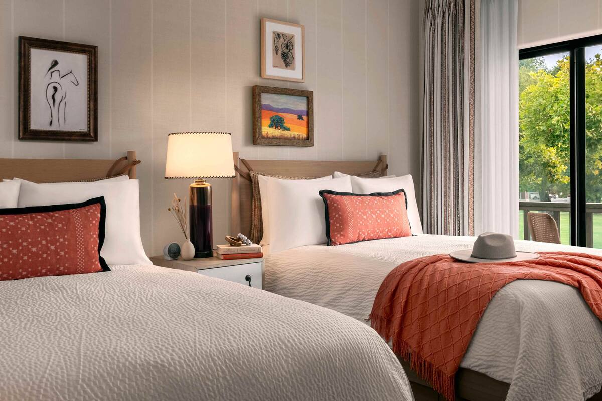 A hotel room with two beds, a nightstand, a lamp, framed artwork, and a view of greenery through the window.