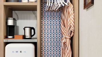 A compact kitchenette with a white refrigerator, coffee maker, and kettle. There are two potted plants on the top shelf and a striped robe hanging on a rack.