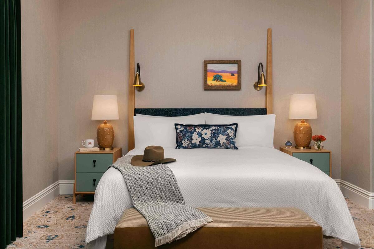 A neatly made bed with a white and blue floral pillow, a brown hat on the bed, and a blanket draped over the side. There are two bedside tables with lamps and decorative vases.