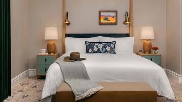 A neatly made bed with a white and blue floral pillow, a brown hat on the bed, and a blanket draped over the side. There are two bedside tables with lamps and decorative vases.