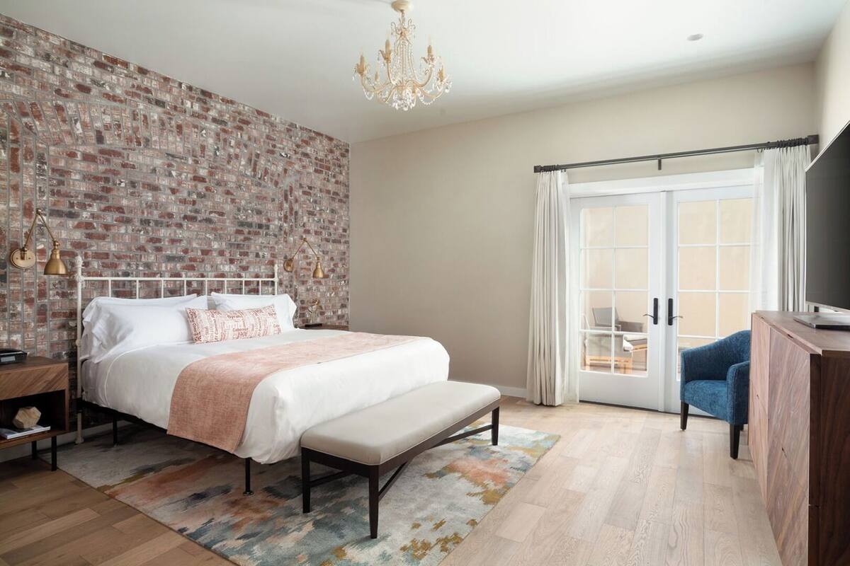 A bedroom with a brick wall, a bed with a bench, a desk with a TV, and a view through a sliding door.