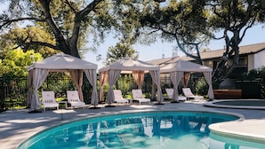A swimming pool with lounge chairs and canopies, surrounded by trees and a building.