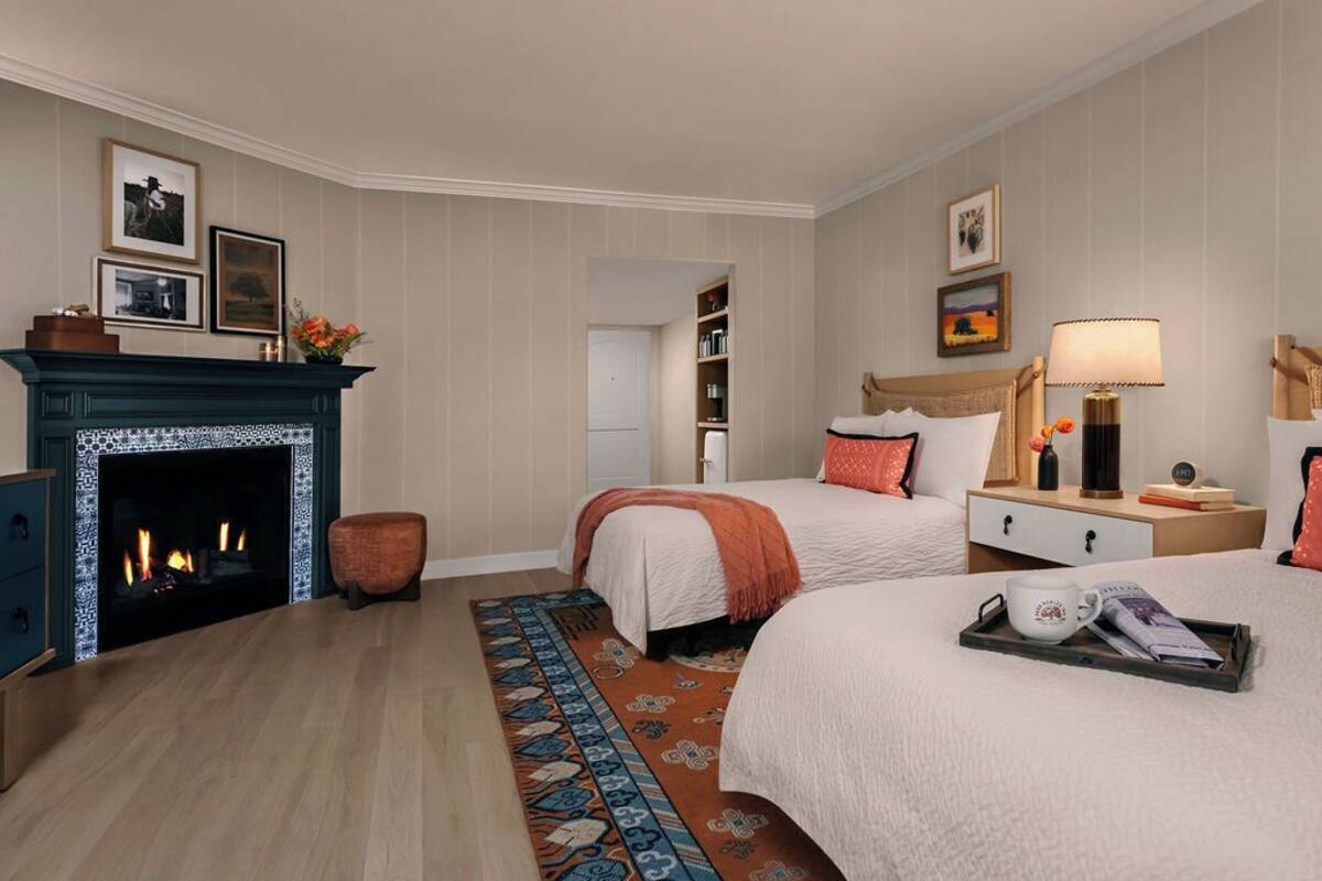 A hotel room with two beds, a fireplace, a nightstand, a lamp, and a tray with a mug and magazine.