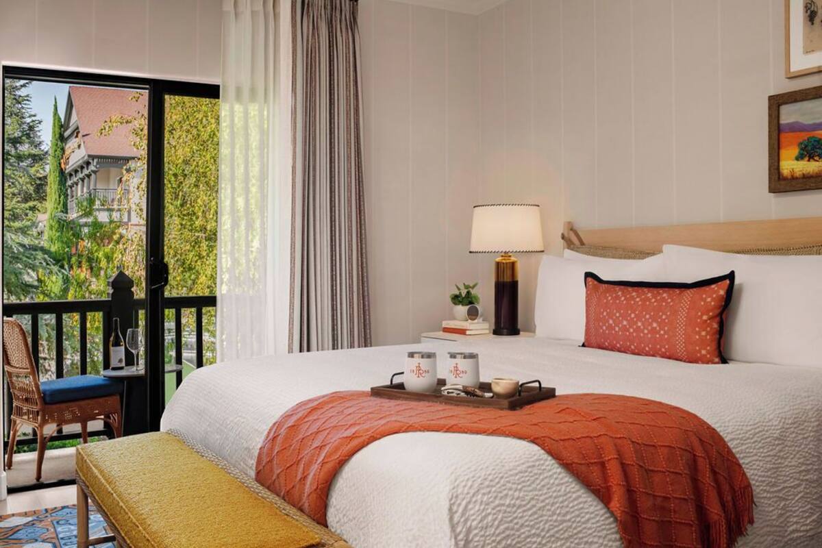 A hotel room with a large bed, a bedside table with a lamp, a small table with candles, and a view of greenery outside.