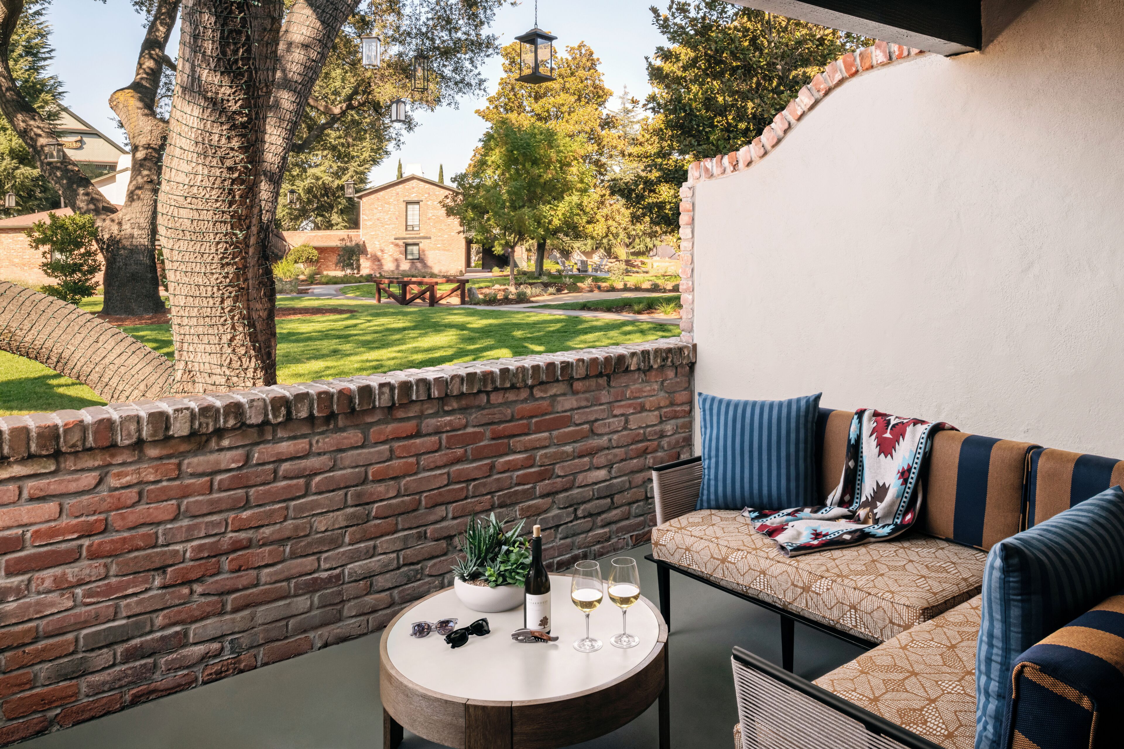 A balcony with a brick wall, a round table with a bottle and glasses, and a sofa with cushions.