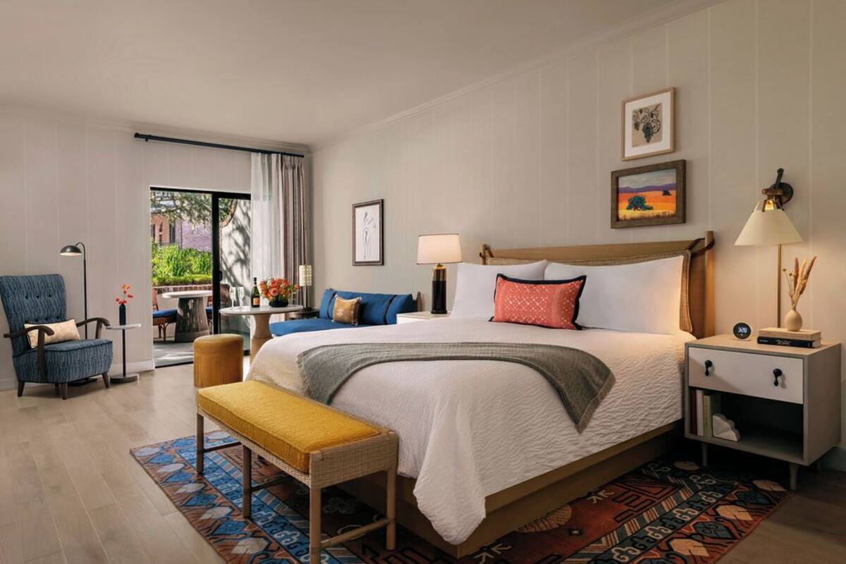 A hotel room with a large bed, a desk, a chair, a bedside table, and a window with curtains.