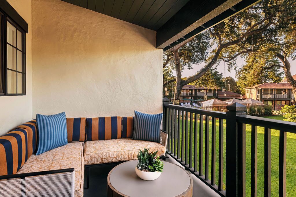 A covered outdoor seating area with a sofa and a round table, overlooking a well-maintained lawn and neighboring buildings.