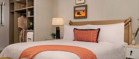 A neatly arranged hotel room with a large bed, a bedside table, a nightstand, a wardrobe, and a small table with a lamp.