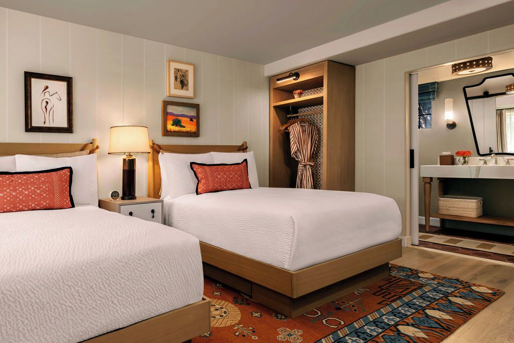 A hotel room with two beds, a nightstand with a lamp, a wardrobe, a mirror, and a small shelf with decorative items.