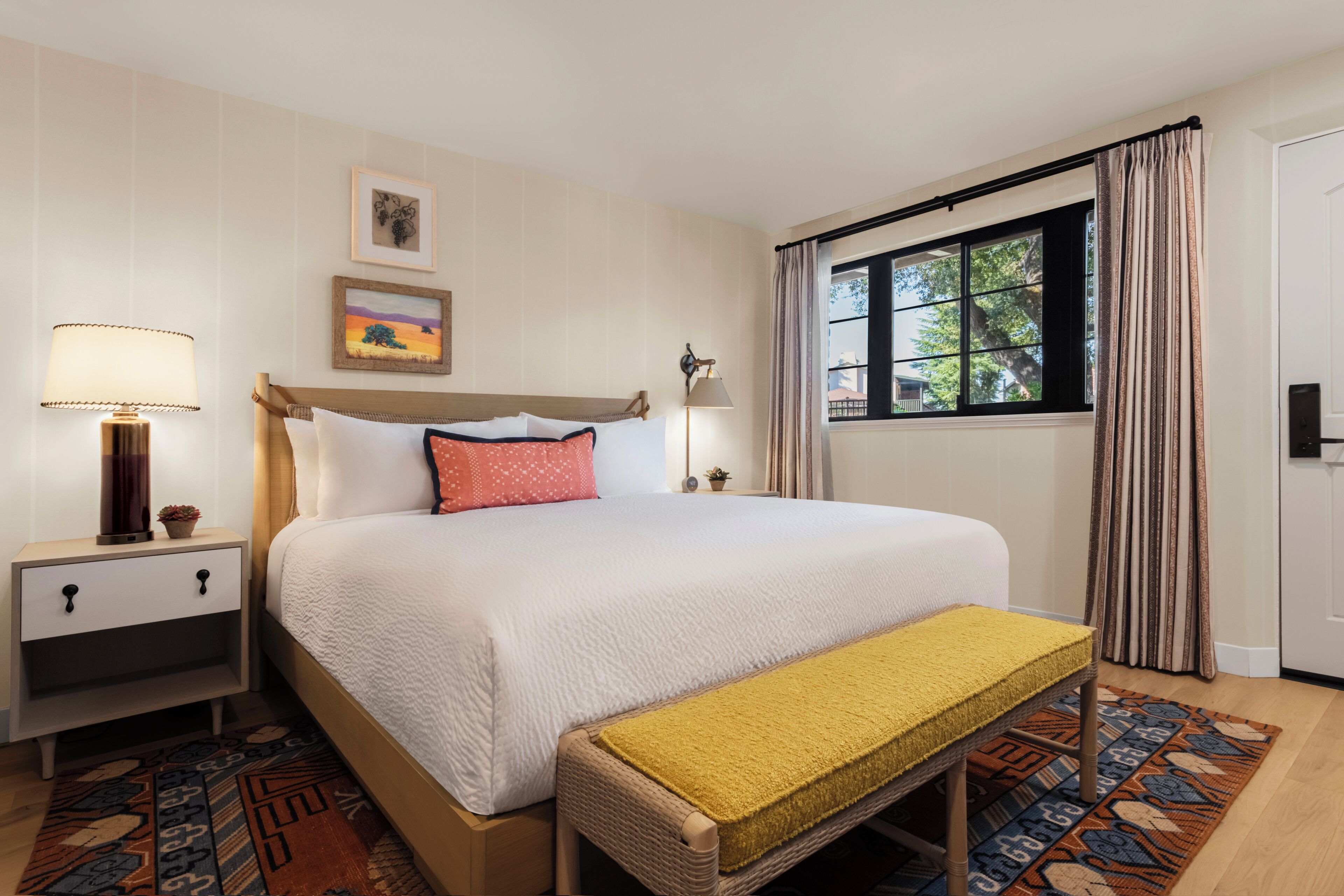 Ein Hotelzimmer mit einem großen Bett, einem Nachttisch mit Lampe, einem kleinen Tisch mit Pflanze, einem Fenster mit Vorhängen und einem gemusterten Teppich.