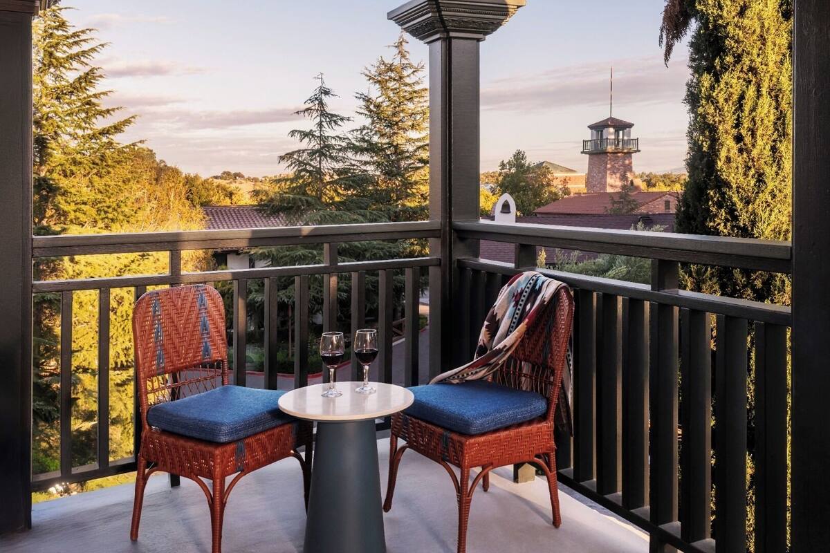 A balcony with wicker chairs and a table, overlooking a scenic view of trees and buildings.