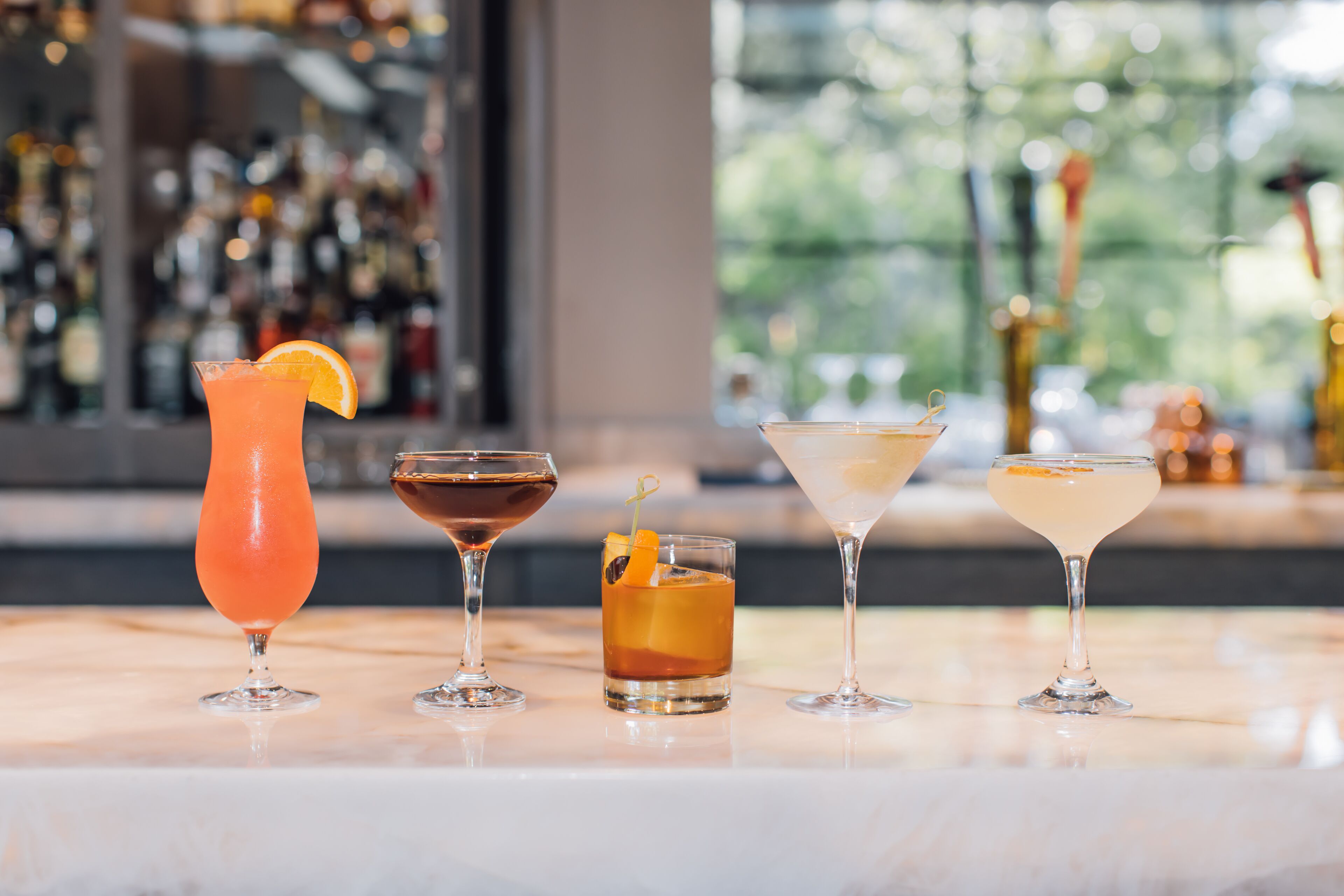 Quatre cocktails différents présentés sur un comptoir de bar en marbre, avec un arrière-plan flou composé de bouteilles et d’une fenêtre.