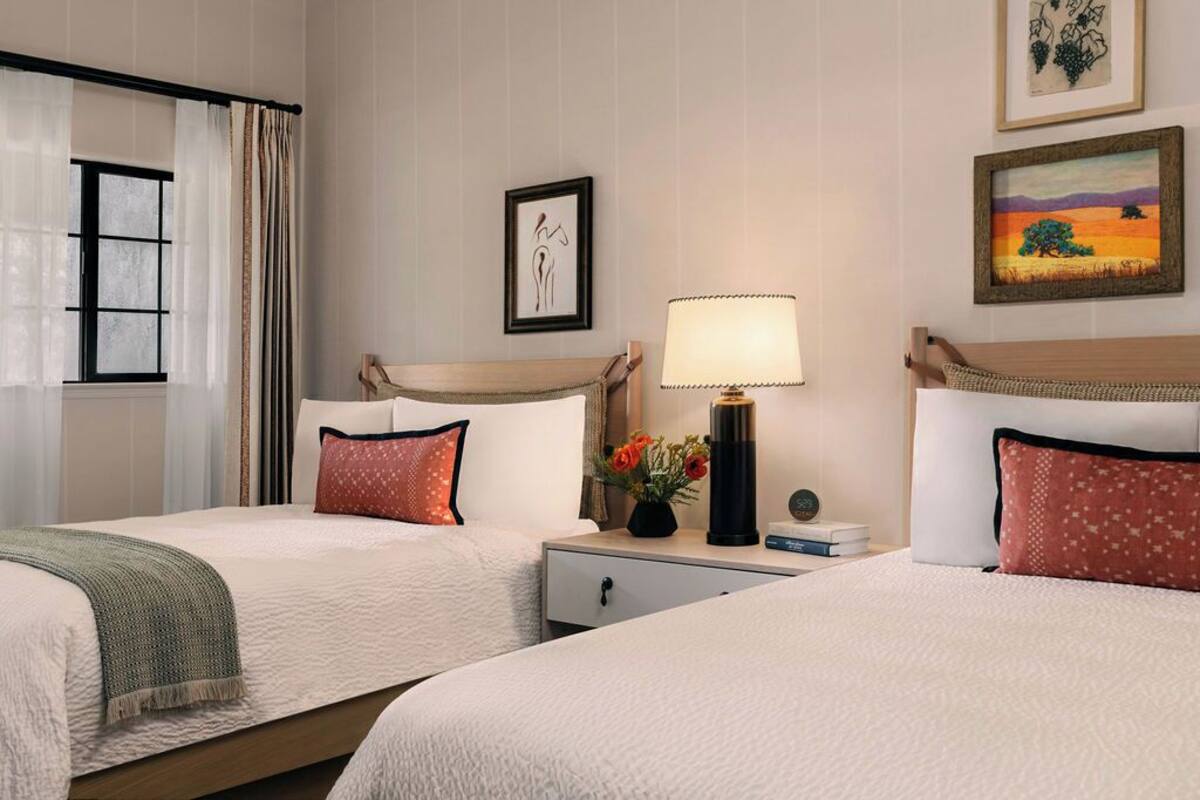 A hotel room with two beds, a nightstand with a lamp, a framed artwork, and a window with curtains.