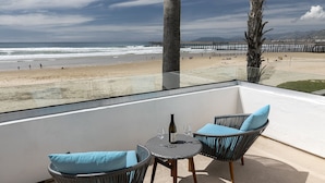 Un balcón con sillas de mimbre y una mesa, con vistas a una playa que tiene un muelle y montañas al fondo.