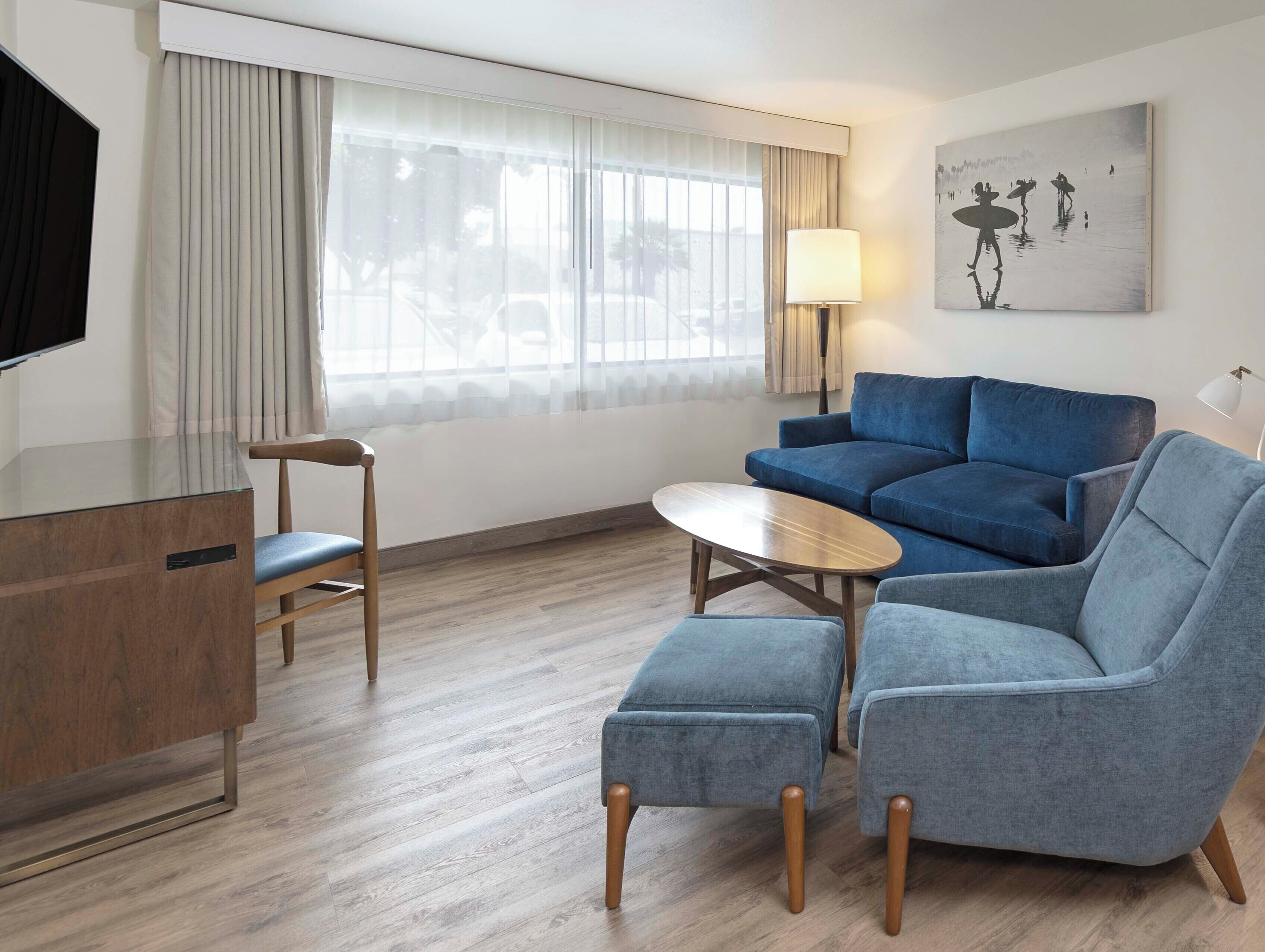 A modern hotel room with a sofa, armchair, ottoman, a small round table, a TV, and a painting on the wall.