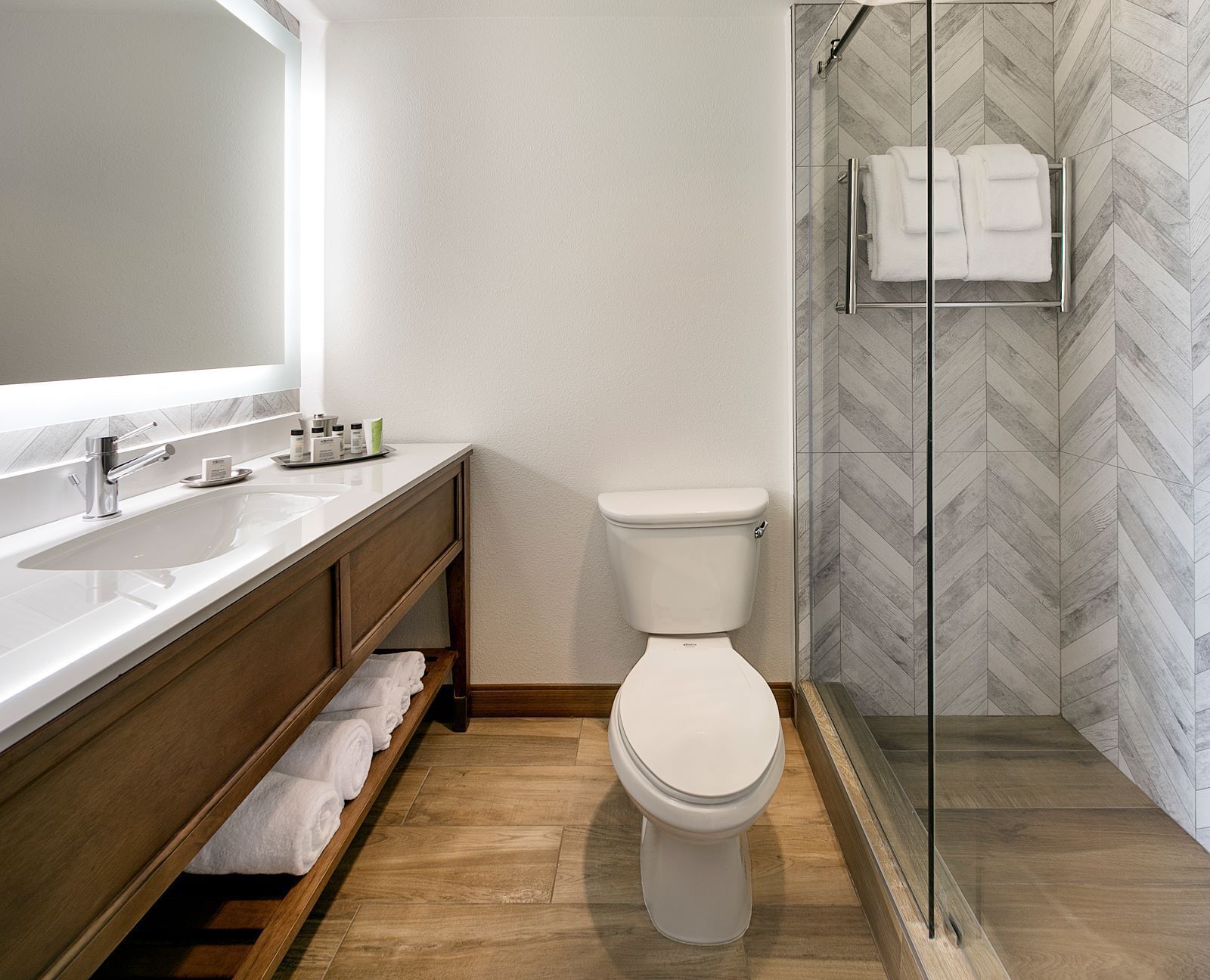 Un baño moderno con inodoro blanco, un espejo grande, un mueble de tocador de madera y cabina de ducha de vidrio.