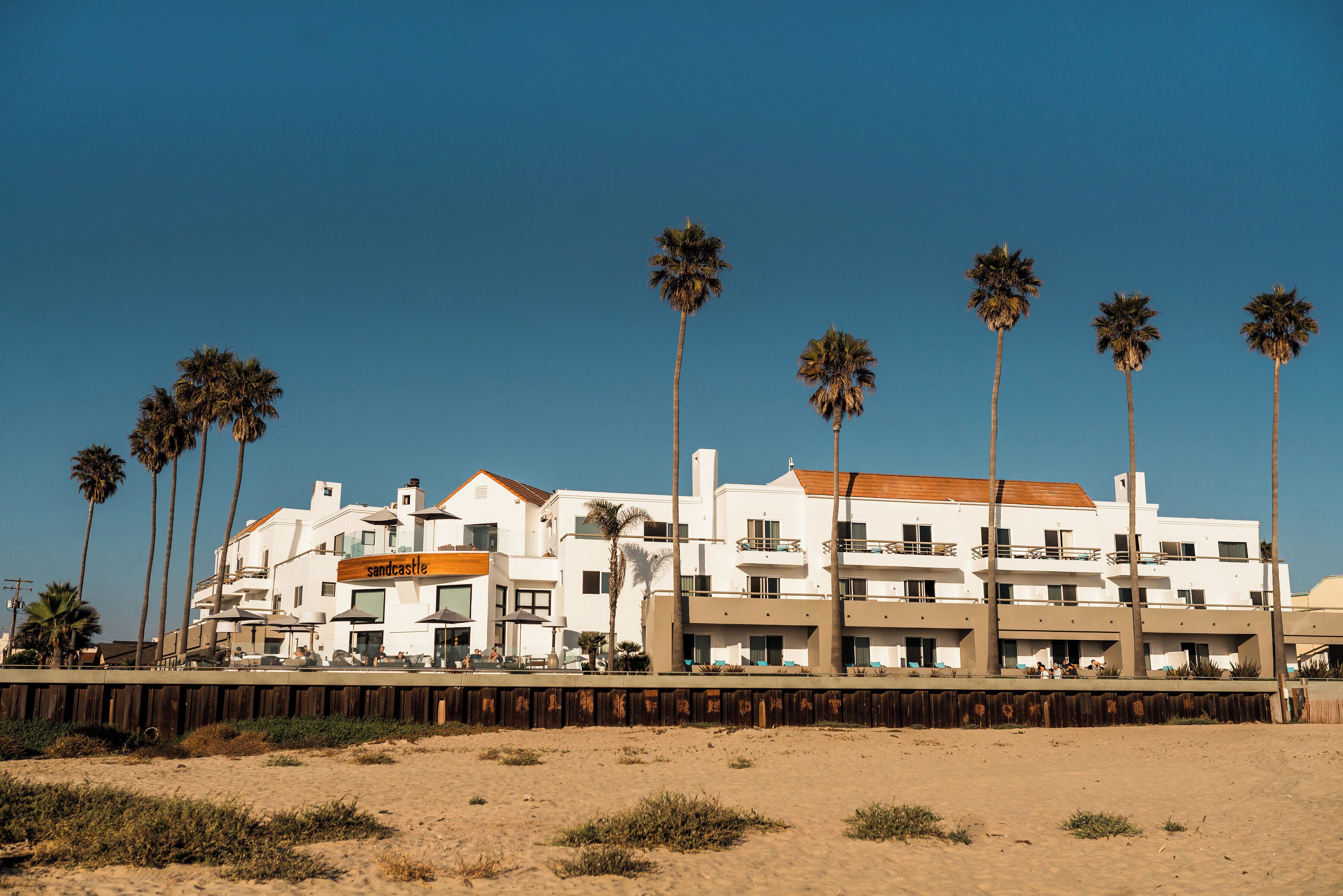 Un hotel frente a la playa con varios pisos, palmeras y una playa arenosa en primer plano.
