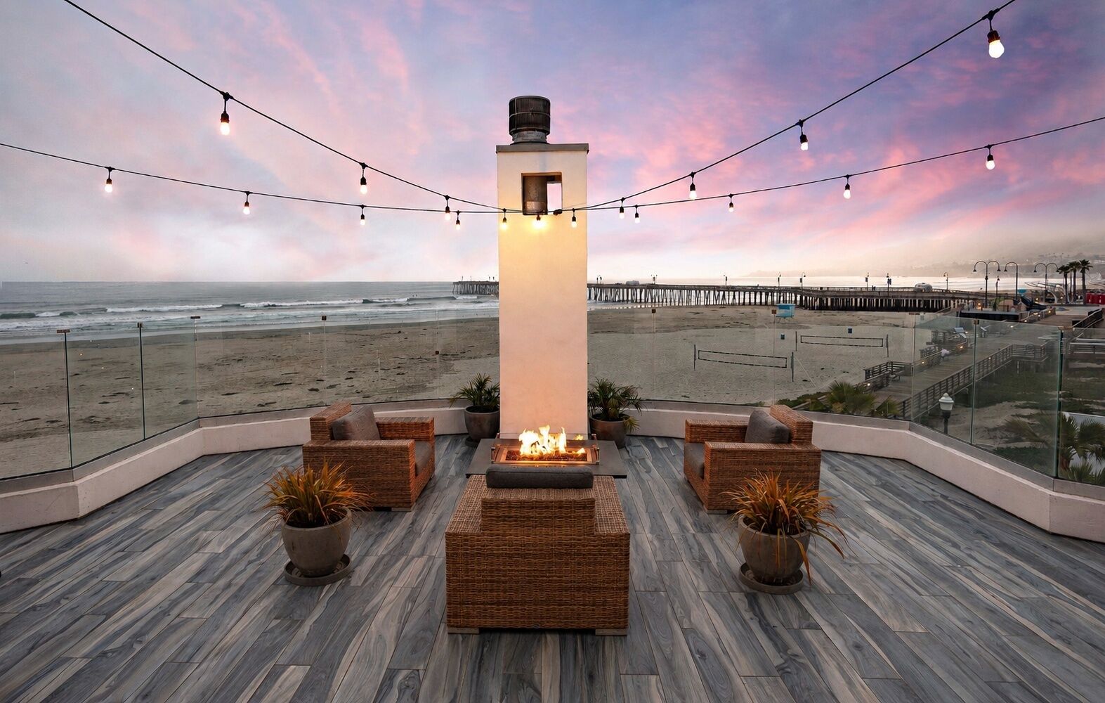 A rooftop lounge with a fire feature, wicker furniture, and a view of the beach and pier.