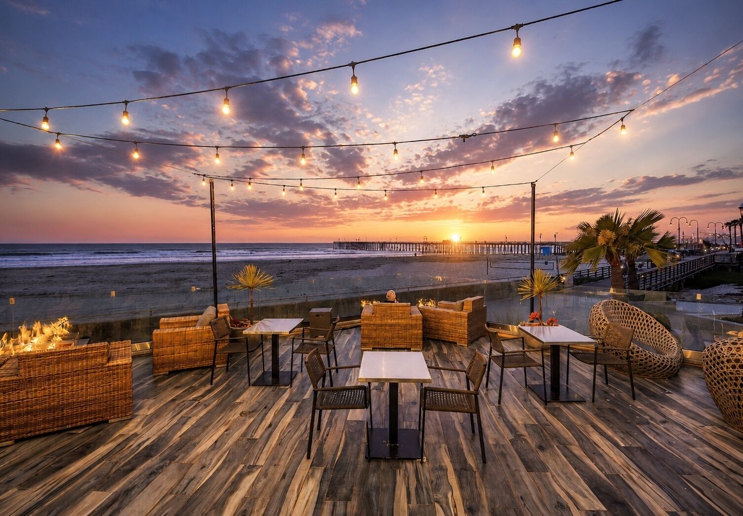An outdoor seating area with wicker furniture and tables, overlooking a beach and a pier at sunset.