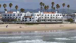 Un hotel frente a la playa con varios balcones, una playa de arena y palmeras.