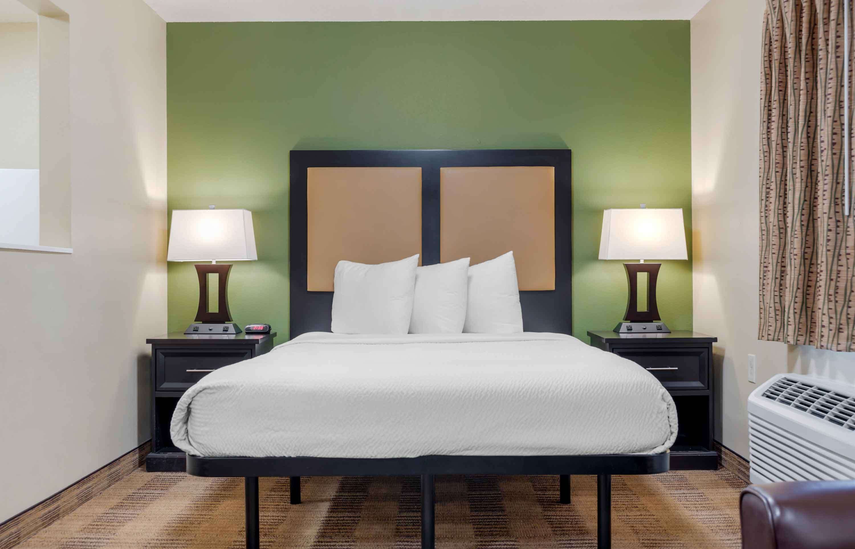 Habitación de hotel con una cama grande, dos mesitas de noche con lámparas, una pared de acento verde y una ventana con cortinas.