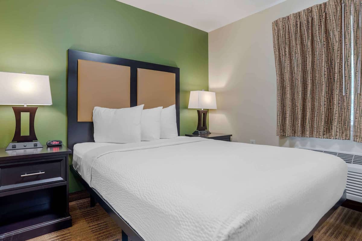 A hotel room with a large bed, two bedside tables with lamps, a nightstand, and a window with curtains.