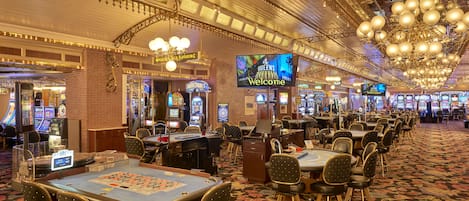 A casino interior with multiple gaming tables, slot machines, and a large screen displaying "Welcome" to the "Queens Welcome".