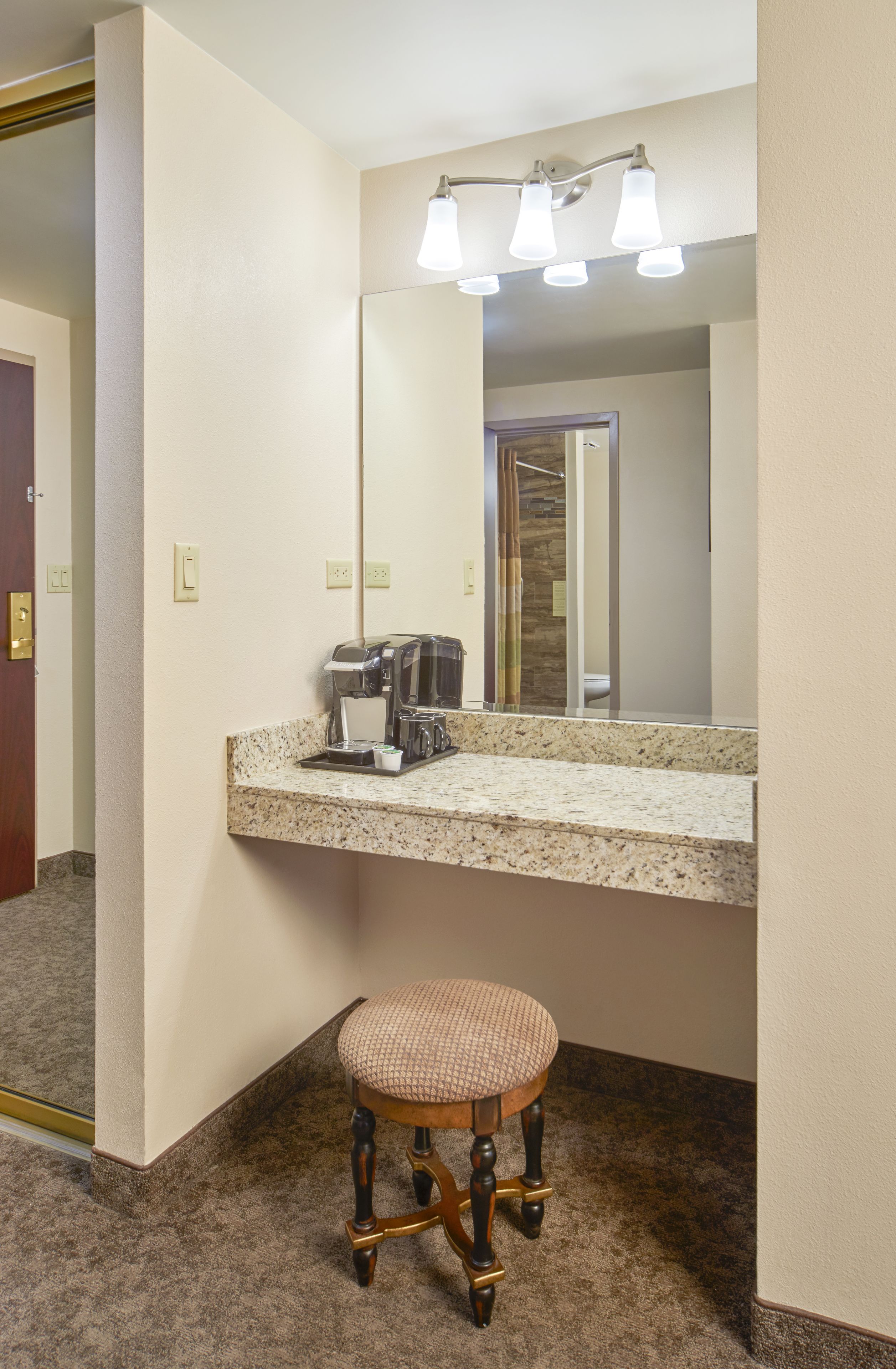 A bathroom with a granite countertop, a coffee maker, a mirror, and a wooden stool.