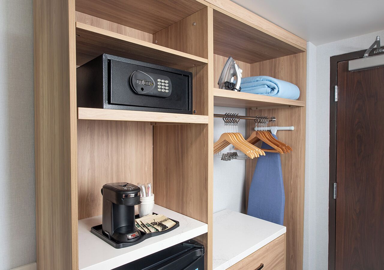 A hotel room with a wooden wardrobe, a coffee maker, a set of keys, and a folded towel on the shelf.