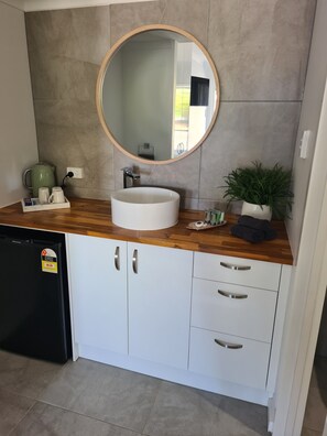A modern bathroom with a round mirror, white sink, and wooden countertop. There is a green plant on the counter and a black dishwasher in the corner.